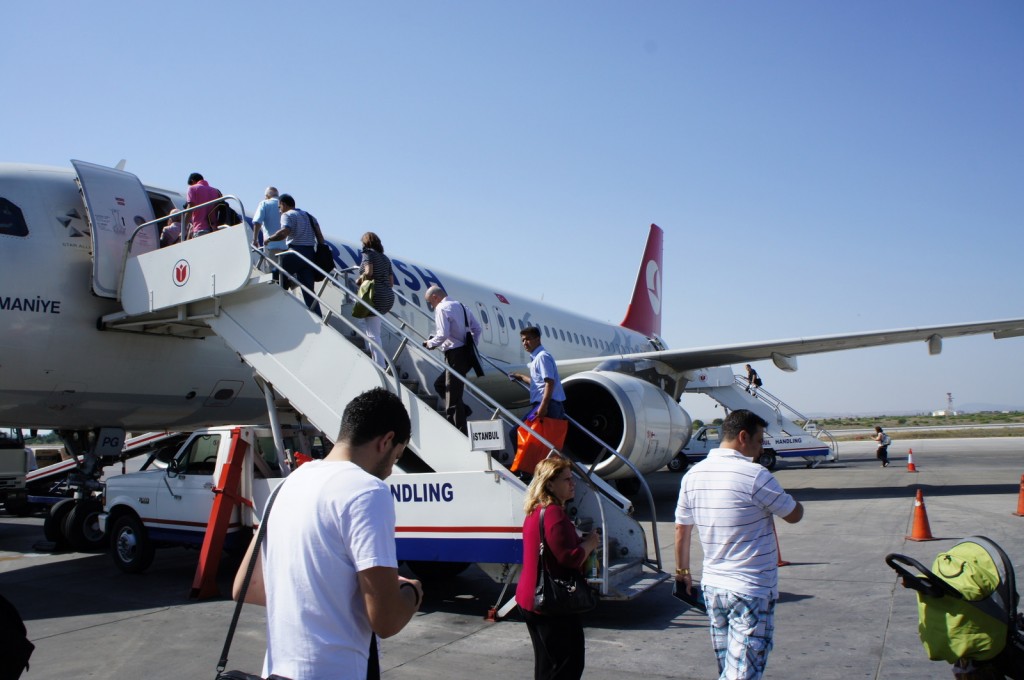 The image shows passengers boarding a Turkish Airlines aircraft via a mobile staircase on the tarmac. The plane is parked on the runway, and several people are seen ascending the stairs to enter the aircraft. The sky is clear and blue, indicating good weather. There are also some ground service vehicles and orange traffic cones visible around the plane.