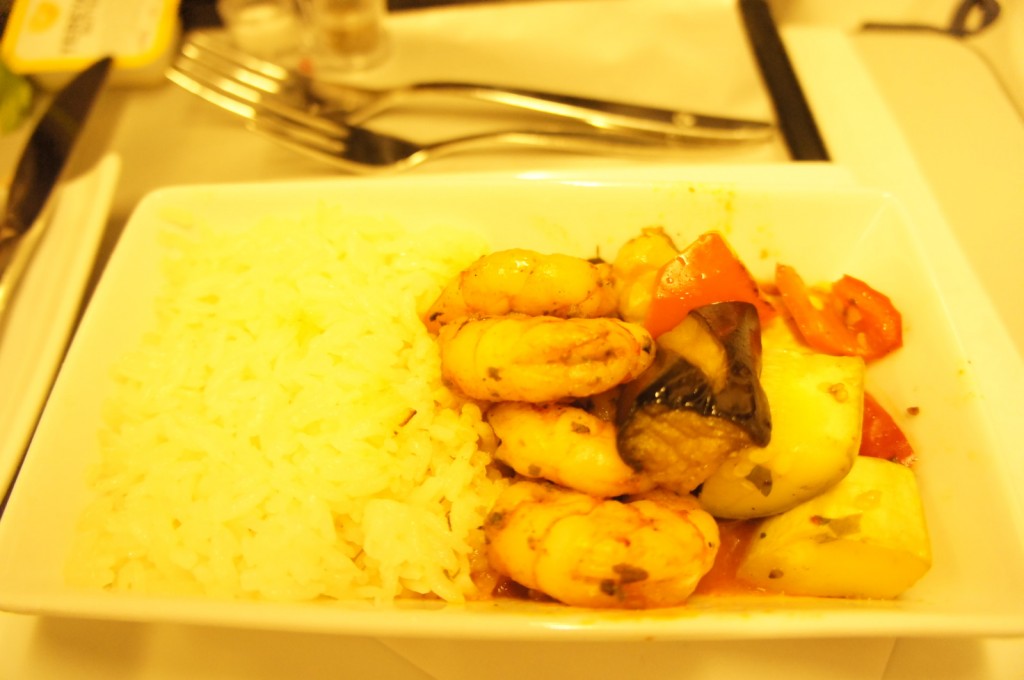A rectangular white dish contains a serving of white rice on the left side and a mix of cooked shrimp, red bell peppers, zucchini, and eggplant on the right side. The dish is placed on a table with a fork and knife in the background.