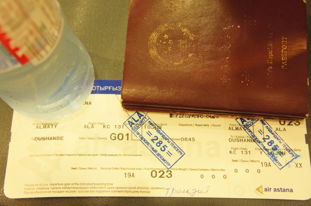 The image shows a close-up of a travel setup including a passport, a boarding pass, and a bottle of water. The passport is partially covering the boarding pass, which is for a flight from Almaty (ALA) to Dushanbe (DYU) on Air Astana, flight number KC 131. The boarding pass has several stamps on it. The bottle of water is placed to the left of the boarding pass and passport.