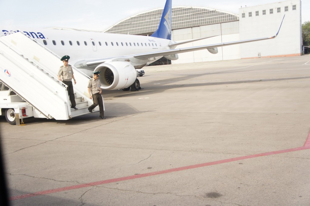 The image shows two uniformed personnel walking down a mobile staircase that is positioned next to a parked airplane on an airport tarmac. The airplane has a white body with blue accents and the word "Avianca" partially visible on the fuselage. The background includes a large hangar and other airport buildings. The scene appears to be taking place in a secure area of the airport.