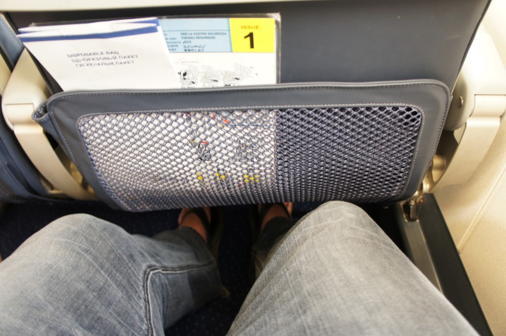 The image shows the view from a passenger's seat on an airplane. The passenger's legs, wearing jeans, are visible in the foreground. The seatback in front of them has a mesh pocket containing a safety information card, a disposable bag, and other literature. The pocket is attached to the back of the seat, which is upholstered in dark material. The floor of the airplane cabin is also visible.