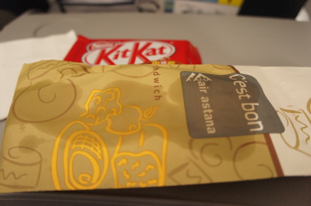 The image shows a close-up of a packaged sandwich with a label that reads "C'est bon" and "Air Astana." The packaging is decorated with yellow illustrations of food items. In the background, there is a red KitKat chocolate bar partially visible. The items are placed on a surface, possibly an airplane tray table.