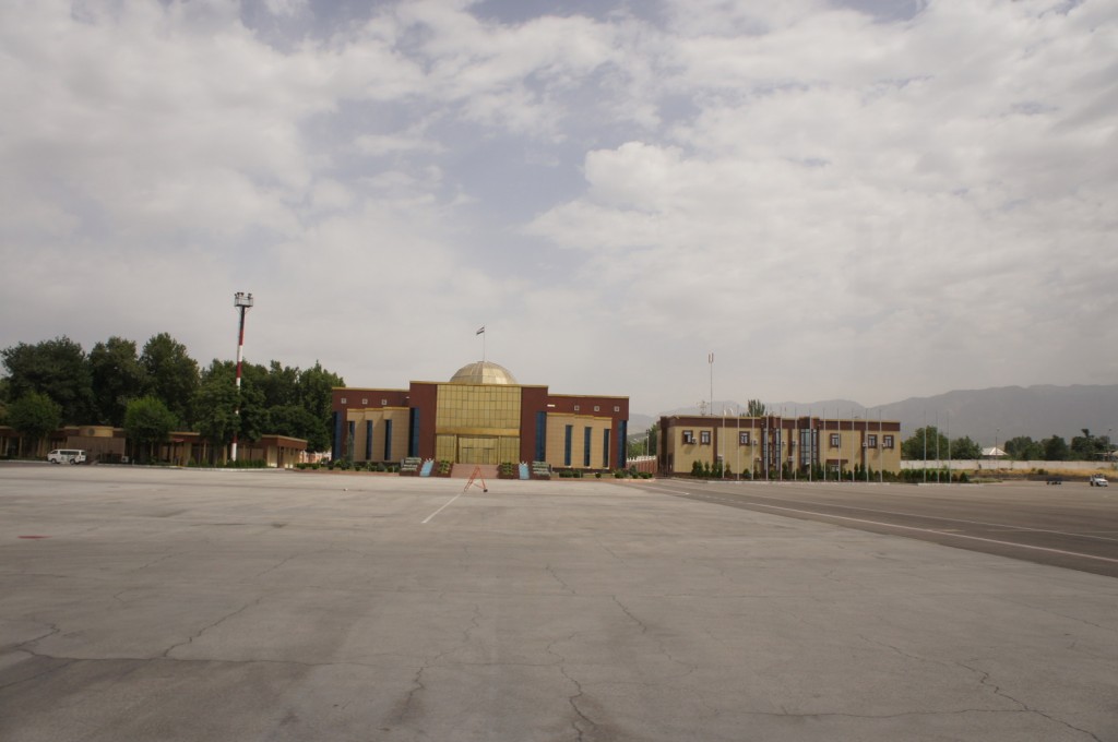 The image shows a large, open concrete area, possibly an airport tarmac or a large parking lot, with a building in the center background. The building has a modern design with a prominent golden dome and large glass windows. There are trees and greenery on the left side of the building, and a row of flagpoles with flags on the right side. The sky is partly cloudy, and there are mountains visible in the distant background.