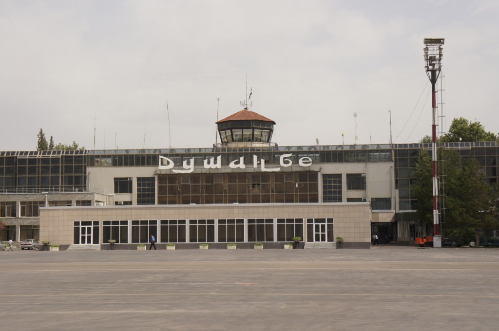 Alt text: "A front view of an airport terminal building with a control tower on the roof. The building has large windows and a sign with Cyrillic letters. There is a tall light pole on the right side and a few trees in the background."