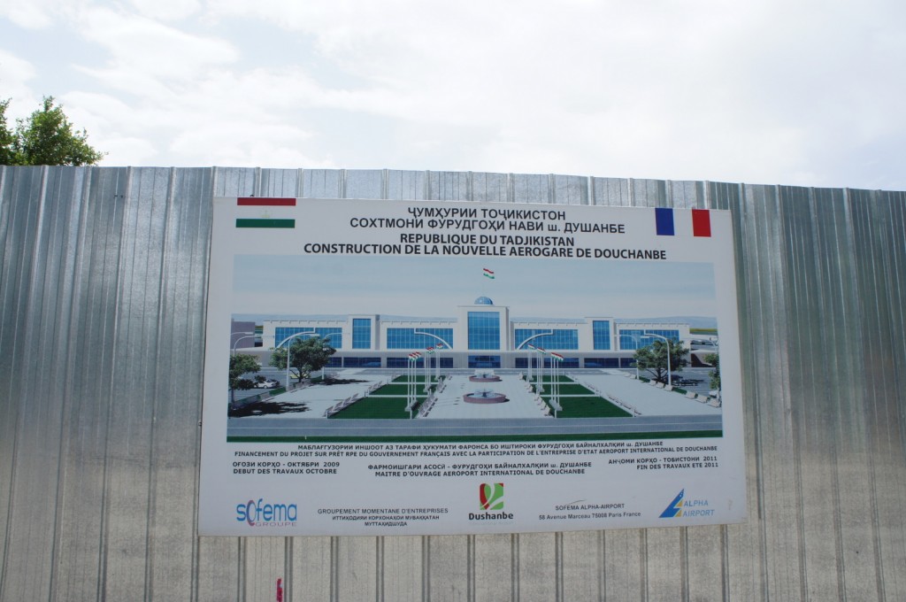 Alt text: A sign on a metal fence displays information about the construction of a new airport terminal in Dushanbe, Tajikistan. The sign includes text in multiple languages, including Tajik, French, and English, and features an illustration of the planned terminal building. The illustration shows a modern structure with a central dome, surrounded by landscaped areas and flagpoles. The logos of Sofema Group, Dushanbe, and Alpha Airport are visible at the bottom of the sign. The sky is partly cloudy, and a tree is visible in the background.