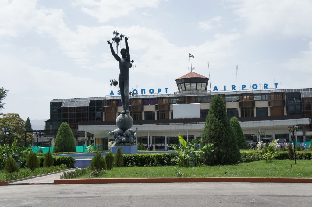The image shows the exterior of an airport building with the word "AIRPORT" written in both Cyrillic and English on the roof. In front of the building, there is a tall statue of a person holding up several objects, possibly representing stars or lights. The statue stands on a pedestal that includes a globe and other decorative elements. The area around the statue is landscaped with green bushes, trees, and a small garden. The sky above is partly cloudy.
