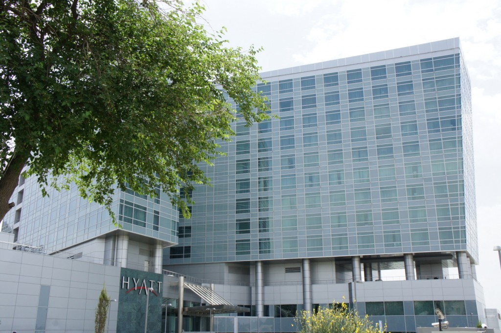 The image shows a modern, multi-story building with a glass facade. The building has a sign that reads "Hyatt" near the entrance. There is a large tree in the foreground, partially obscuring the view of the building. The sky is partly cloudy.