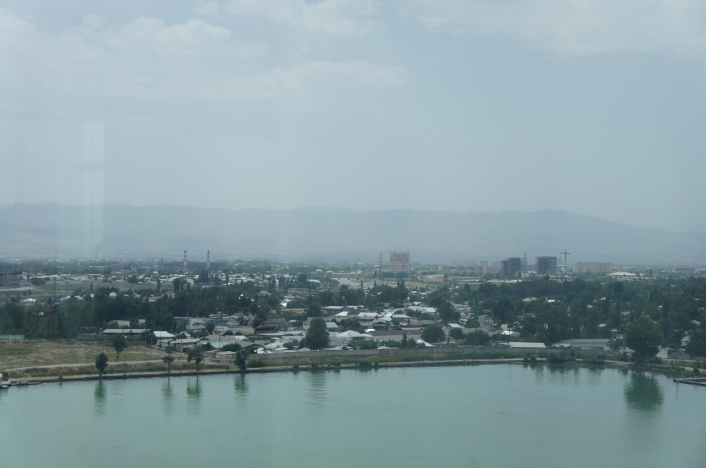 A hazy cityscape with a body of water in the foreground. The city has a mix of low-rise buildings and taller structures under construction, with cranes visible. The background features a range of mountains under a cloudy sky. Trees and greenery are scattered throughout the urban area.