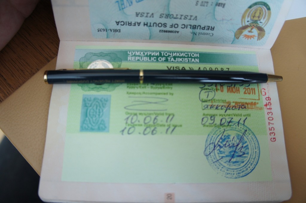 The image shows an open passport displaying a visa for the Republic of Tajikistan. The visa includes various stamps, signatures, and dates, indicating entry and validity periods. A black pen with gold accents is placed horizontally across the open passport. The background includes a wooden surface and part of another document or folder.