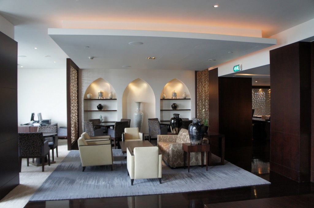 The image shows a modern, elegantly designed lounge area. The space features a mix of contemporary and traditional decor elements. There are several seating arrangements with cushioned chairs and sofas around coffee tables. The walls have built-in shelves with decorative items such as vases and sculptures. The lighting is soft and ambient, creating a cozy atmosphere. There is a desk area with chairs on the left side, and a passageway on the right leading to another room. The floor is covered with a large area rug, and the overall color scheme is neutral with dark wood accents.