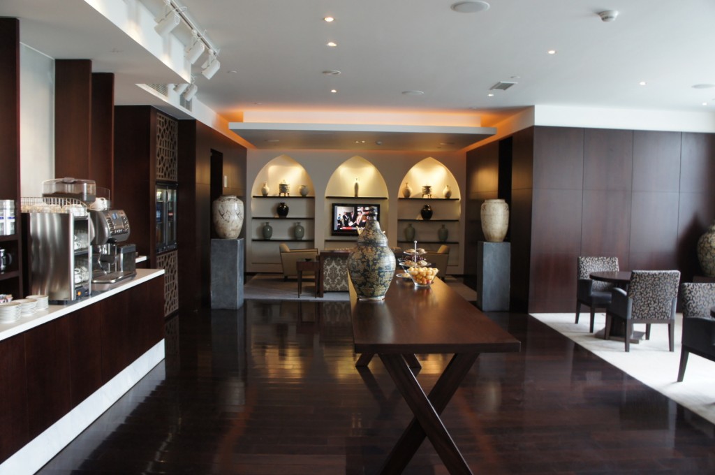 The image shows a modern, stylish lounge area with dark wooden floors and furniture. In the foreground, there is a long wooden table with decorative vases and bowls of fruit. To the left, there is a coffee station with an espresso machine and cups. In the background, there are shelves with various decorative items and a flat-screen TV mounted on the wall. The seating area includes cushioned chairs and small tables. The room is well-lit with recessed lighting and has a contemporary design.