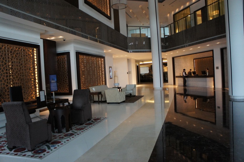 The image shows a modern hotel lobby with a spacious and elegant design. The lobby features a seating area with several chairs and small tables arranged on a decorative rug. The walls have intricate lattice designs, and there is a reception desk in the background where two staff members are assisting guests. The floor is a combination of light and dark polished tiles, and the area is well-lit with both natural and artificial lighting. The overall ambiance is sophisticated and welcoming.