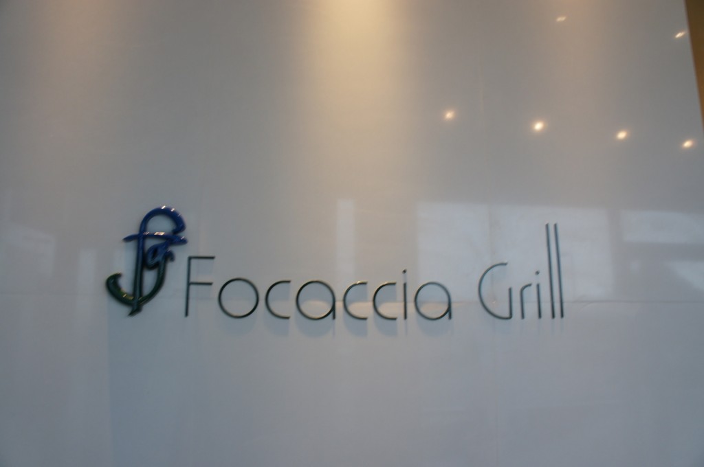 The image shows a wall with the text "Focaccia Grill" written on it. There is also a stylized logo to the left of the text, which appears to be a combination of the letters "F" and "G" in blue and green colors. The background is a smooth, light-colored surface, and there are some lights reflecting off the wall.