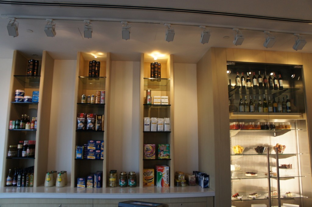 The image shows a well-organized display of various food and beverage items on shelves in a store or caf&eacute;. The shelves are built into the wall and are illuminated by spotlights from above. The items on the shelves include canned goods, boxed foods, jars, bottles, and other packaged products. On the right side, there is a glass cabinet with a selection of wines on the top shelf and desserts or other food items on the lower shelves. The overall setting appears clean and modern.