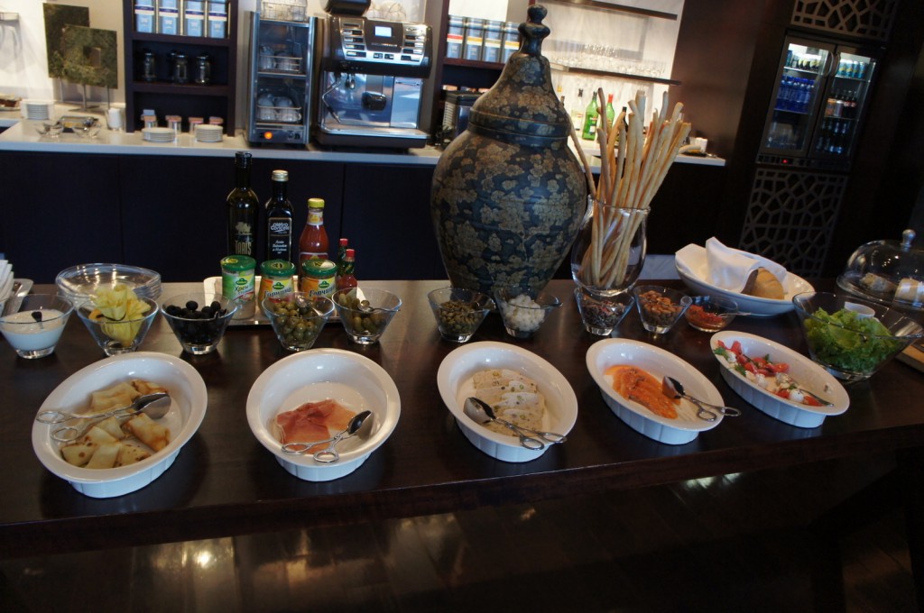 The image shows a buffet setup with various food items displayed on a table. There are several white dishes containing different types of food, including what appears to be grilled vegetables, slices of meat, and salads. In addition, there are bowls with ingredients such as olives, capers, and pineapple chunks. There are also bottles of olive oil, sauces, and canned goods. A large decorative urn and a container with breadsticks are placed in the center of the table. In the background, there is a counter with coffee machines, shelves with jars, and a refrigerator with beverages.
