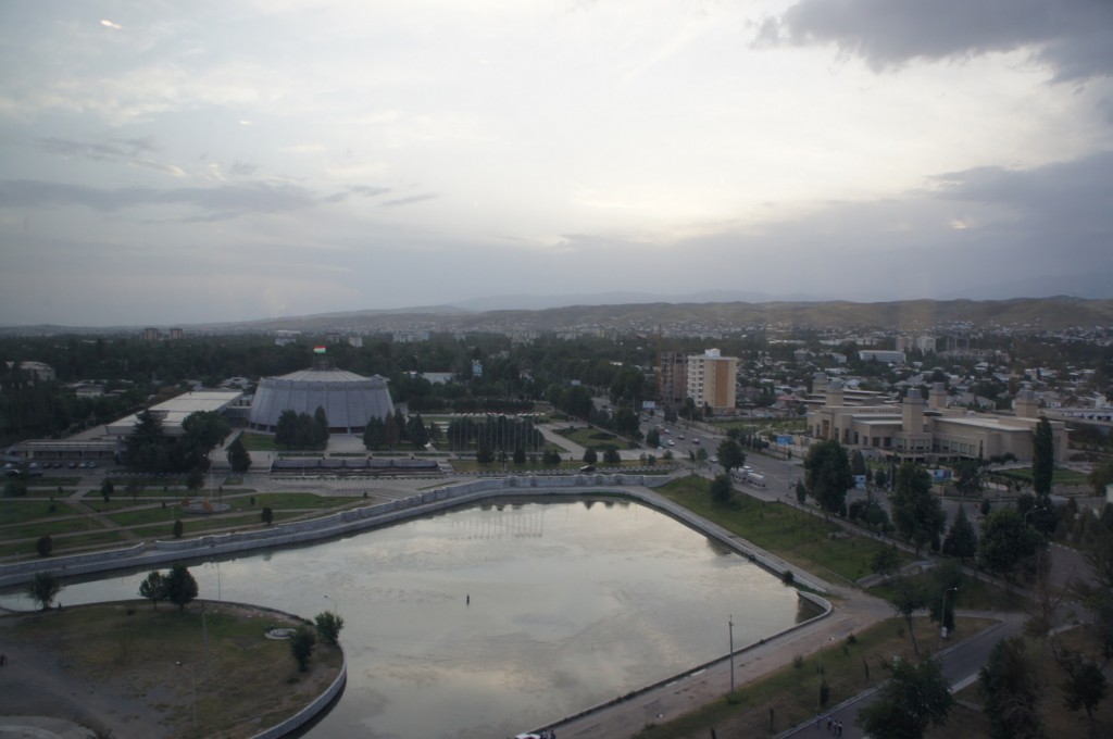 A cityscape view at dusk, featuring a large, hexagonal building with a flag on top, surrounded by trees and smaller buildings. In the foreground, there is a body of water with a curved shoreline, and a road running alongside it. The background shows a sprawling urban area with various buildings and a hilly landscape under a cloudy sky.