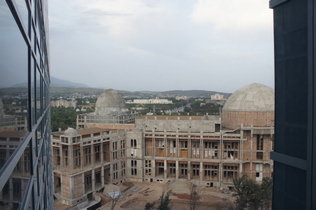 The image shows a large, unfinished building with a dome-shaped roof, viewed from a higher vantage point. The building appears to be under construction, with exposed brickwork and scaffolding visible. In the background, there are more buildings and a hilly landscape. The sky is overcast, and the reflection of the building is visible on the glass facade of an adjacent structure.