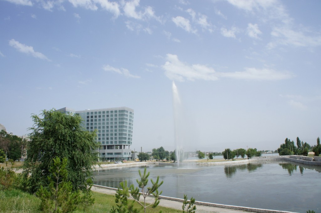 The image shows a large, modern building with many windows situated near a body of water. In the water, there is a tall fountain spraying water high into the air. The scene is set on a clear day with a blue sky and some scattered clouds. There are trees and greenery in the foreground and background, adding a natural element to the urban setting.