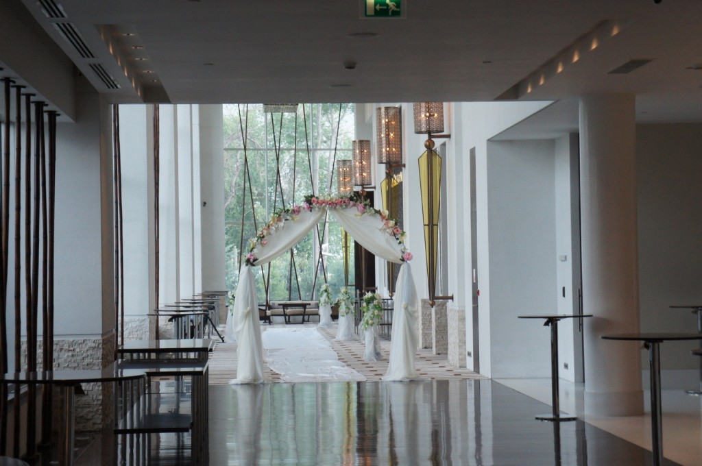 A spacious, modern hallway with a high ceiling and large windows letting in natural light. The hallway features a decorative archway draped with white fabric and adorned with pink and white flowers, suggesting a wedding or special event setup. There are tables and chairs along the sides, and the floor is shiny and reflective. Large lantern-style lights hang from the ceiling, and there is an exit sign visible at the top. The overall ambiance is elegant and serene.