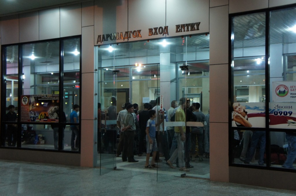 The image shows the entrance of a building with glass doors and windows. Above the entrance, there are signs in three languages: Cyrillic, another script, and English, all indicating "ENTRY." Inside, a group of people is gathered, some standing and some walking. The interior is well-lit, and there are posters and advertisements visible through the windows on either side of the entrance. The scene appears to be in the evening or night, given the artificial lighting.