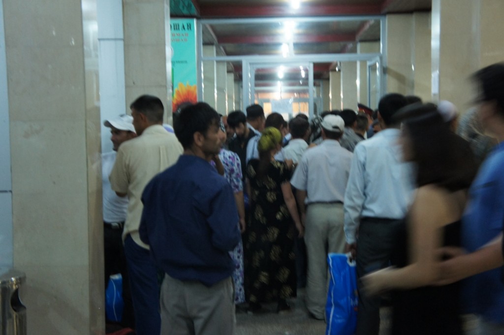 A large group of people is gathered in a hallway, possibly waiting in line. The hallway is indoors with beige walls and a ceiling with red beams. Some people are holding blue bags, and there is a mix of men and women, some wearing hats. The atmosphere appears crowded and busy.