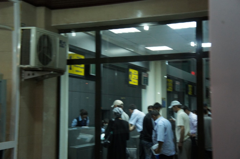 The image shows a group of people inside a building, possibly a bank or a service center. They are standing in line or gathered around a counter where a person behind the counter appears to be assisting them. The room has fluorescent lighting and there are signs with text on the walls. An air conditioning unit is mounted on the wall to the left. The scene is viewed through a glass window or door.
