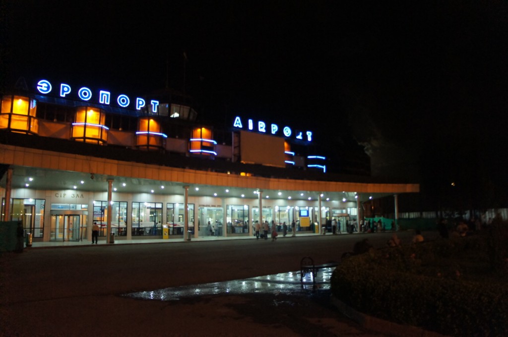 The image shows the exterior of an airport terminal at night. The building is illuminated with bright lights, and the word "АЭРОПОРТ" (which means "airport" in Russian) is prominently displayed in neon letters on the upper part of the structure. The entrance area is well-lit, and there are people visible near the entrance, possibly passengers or staff. The surrounding area appears to be wet, reflecting the lights from the building.
