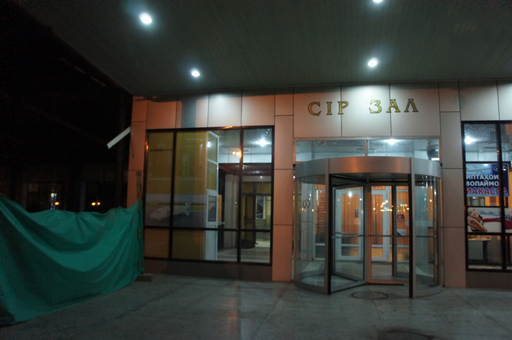 The image shows the entrance to a building at night. The entrance features a revolving glass door and large windows. Above the door, there is a sign with Cyrillic text that reads "CIP ЗАЛ." The area is illuminated by overhead lights, and there is a green tarp or covering to the left side of the entrance. Inside, the building appears to be well-lit, and there are some posters or advertisements visible through the windows on the right side.