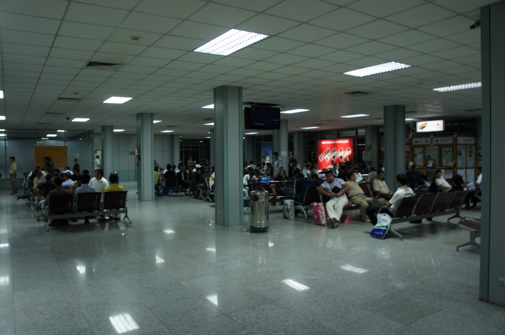 The image shows a large indoor waiting area, likely an airport terminal or a bus station. The space is filled with rows of seating, and many people are sitting and waiting. The ceiling has a grid pattern with fluorescent lights. There are several pillars throughout the area, and a few trash bins are visible. Some people have bags and luggage with them. In the background, there are advertisements and signs on the walls. The overall atmosphere appears to be calm and orderly.