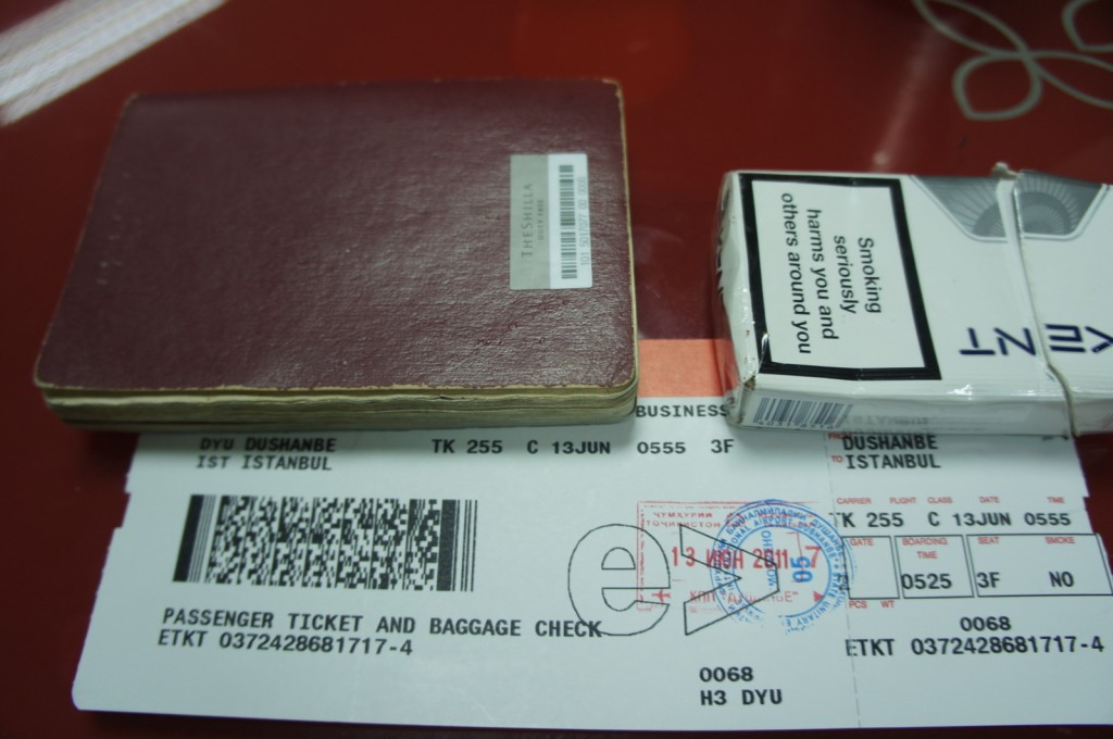 The image shows a red passport, a pack of Kent cigarettes, and a boarding pass. The boarding pass is for a flight from Dushanbe to Istanbul on Turkish Airlines, flight number TK 255, departing on June 13th at 05:55. The seat number is 3F, and the class is business. The boarding pass also has a barcode and several stamps on it.