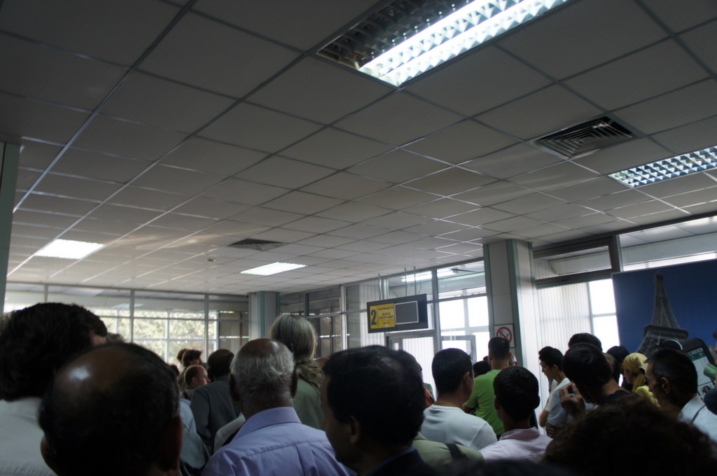 A large group of people is gathered in what appears to be an indoor waiting area or queue. The ceiling has fluorescent lighting, and there are windows along the walls letting in natural light. A sign with the number "2" and some text is visible in the background, along with a television screen. The people are standing closely together, facing towards the sign and screen. The atmosphere suggests a busy or crowded environment, possibly at an airport, office, or service center.