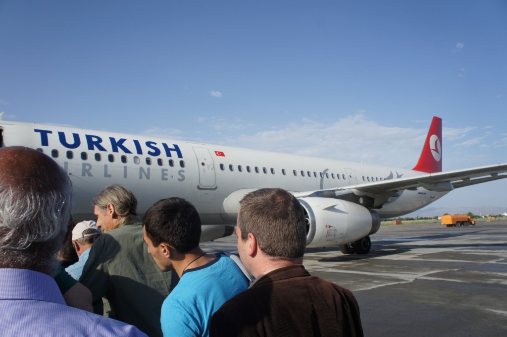 A group of people is seen boarding a Turkish Airlines airplane on a sunny day. The aircraft is parked on the tarmac, and the airline's logo and name are visible on the side of the plane. The sky is clear with a few clouds, and there is some ground equipment in the background.