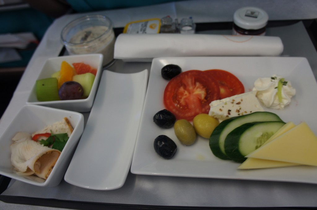 The image shows a meal served on a tray, likely on an airplane. The meal includes a plate with sliced tomatoes, cucumbers, cheese, olives, and a dollop of cream cheese or similar spread. There are also two small bowls, one containing a variety of fresh fruit pieces (including grapes, melon, and citrus) and the other containing a wrap or roll with a filling, possibly turkey or chicken, garnished with a sprig of parsley. Additionally, there is a small container of what appears to be a spread or dip, a rolled napkin, and a small jar of jam or preserve.