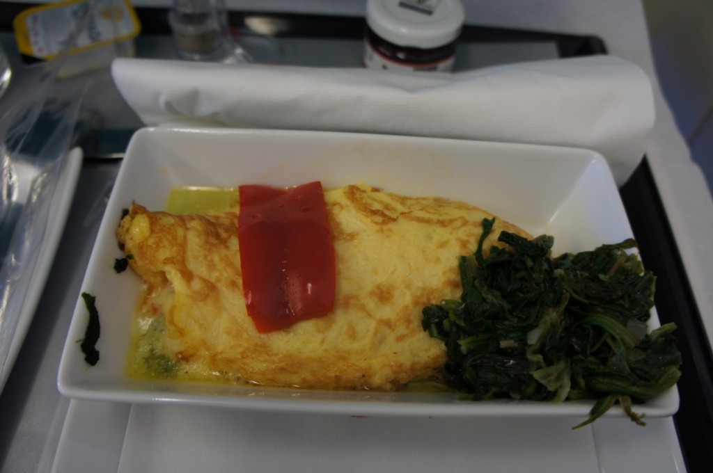 A rectangular white dish contains an omelette topped with a strip of red bell pepper. On the side of the omelette, there is a serving of cooked greens, possibly spinach. The dish is placed on a tray with a white napkin and a small jar in the background.