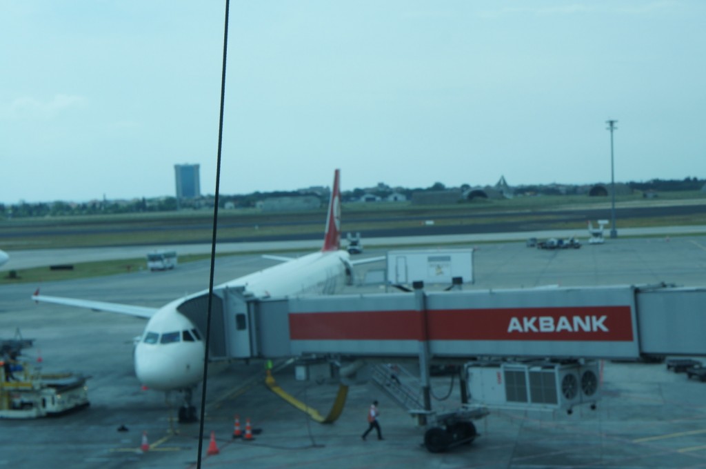 The image shows an airplane parked at an airport gate. The jet bridge, labeled with "AKBANK," is connected to the aircraft. The scene includes airport ground equipment and personnel near the plane. The background features the runway and some distant buildings under a clear sky.