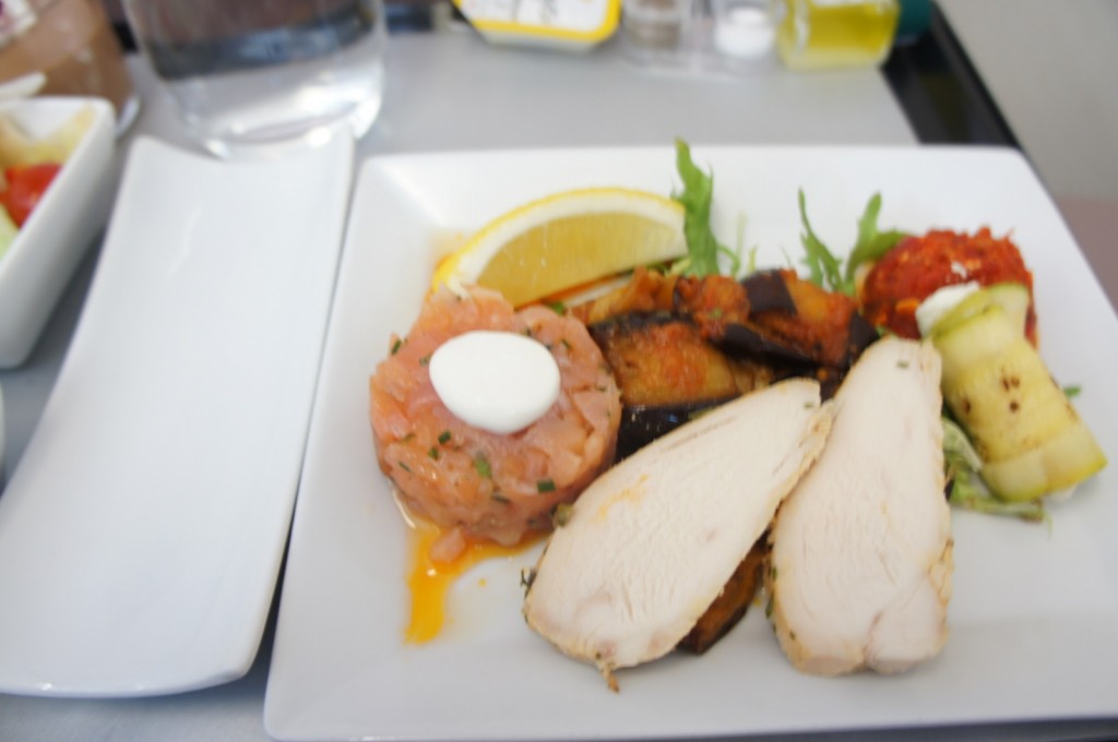 A white square plate with a variety of foods, including slices of cooked chicken breast, a small portion of salmon tartare topped with a dollop of white sauce, grilled eggplant, a lemon wedge, and a small serving of what appears to be a red pepper or tomato-based dish. There is also a piece of rolled zucchini. In the background, there is a glass of water and some condiments.