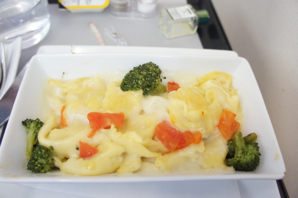 A rectangular white dish contains a serving of cheese tortellini pasta with a creamy sauce, garnished with pieces of broccoli and diced red bell peppers. The dish is placed on a tray, with a glass of water and some condiments visible in the background.