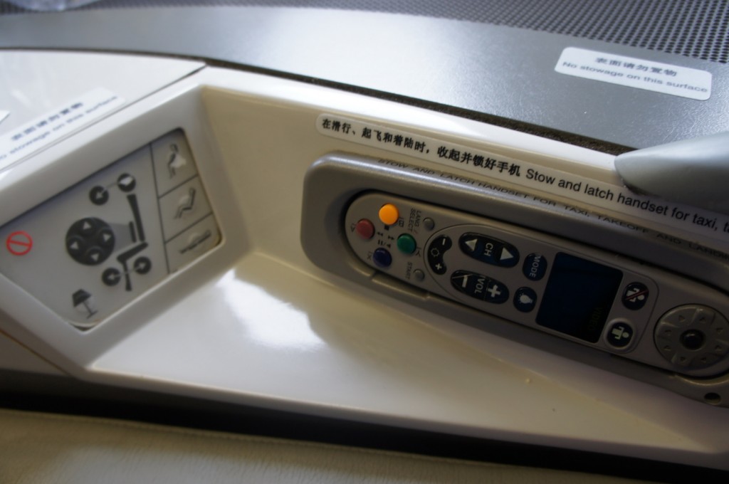 The image shows a control panel and a remote control handset, likely from an airplane seat. The control panel has various buttons for adjusting the seat position and other functions, while the remote control has buttons for in-flight entertainment. There are instructions in both English and Chinese, including a notice that reads "Stow and latch handset for taxi, takeoff, and landing."