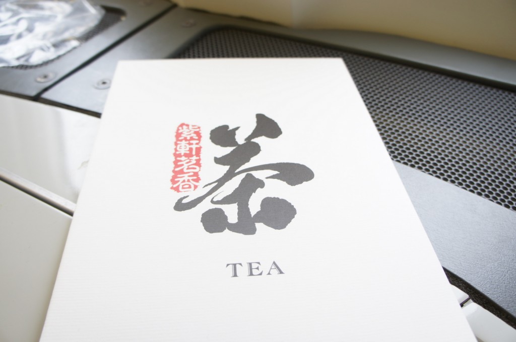 The image shows a white card or menu with the word "TEA" written in English at the bottom. Above the English text, there is a large black Chinese character, and to the left of it, there is a vertical red stamp with additional Chinese characters. The card is placed on a surface with a metallic, perforated texture in the background.