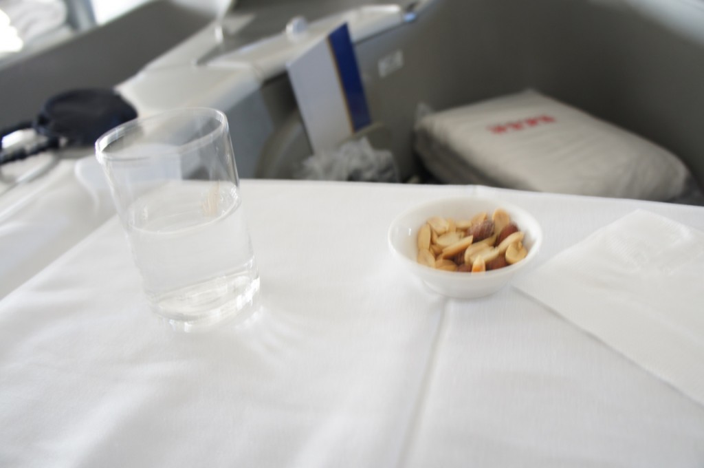 The image shows a close-up of a table with a white tablecloth. On the table, there is a clear glass of water and a small white bowl containing mixed nuts. There is also a white napkin next to the bowl. In the background, there is a blurred view of what appears to be an airplane seat and a pillow.
