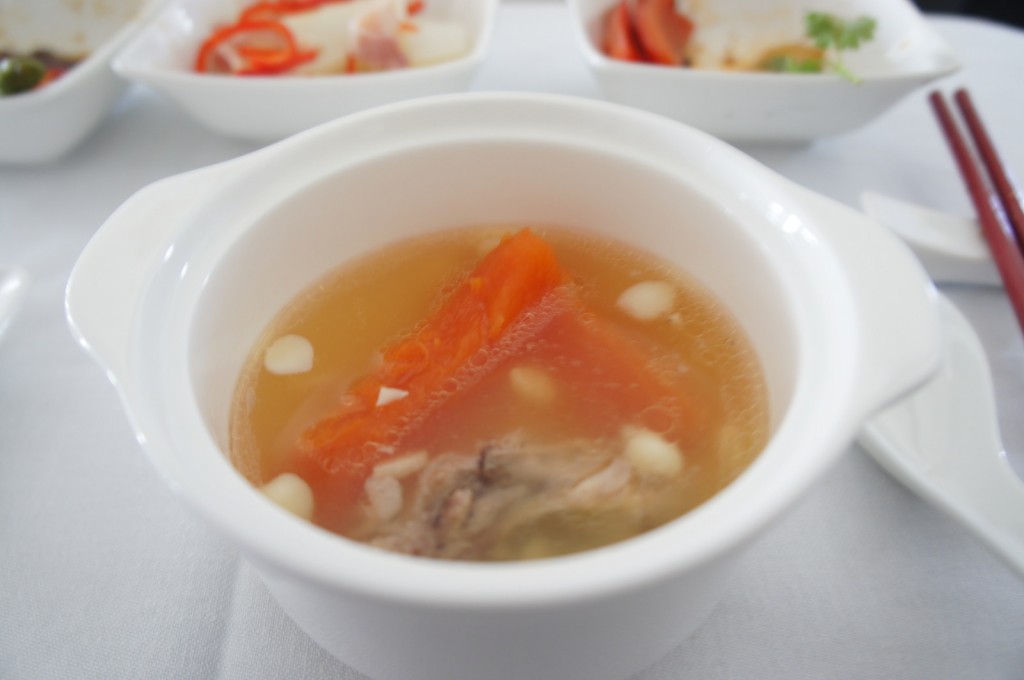 The image shows a bowl of clear soup with visible ingredients including a piece of orange carrot, some small white beans or seeds, and a piece of meat. The bowl is white and has handles on both sides. In the background, there are other dishes in white bowls, and a pair of red chopsticks and a white spoon are placed on the table. The tablecloth is white.