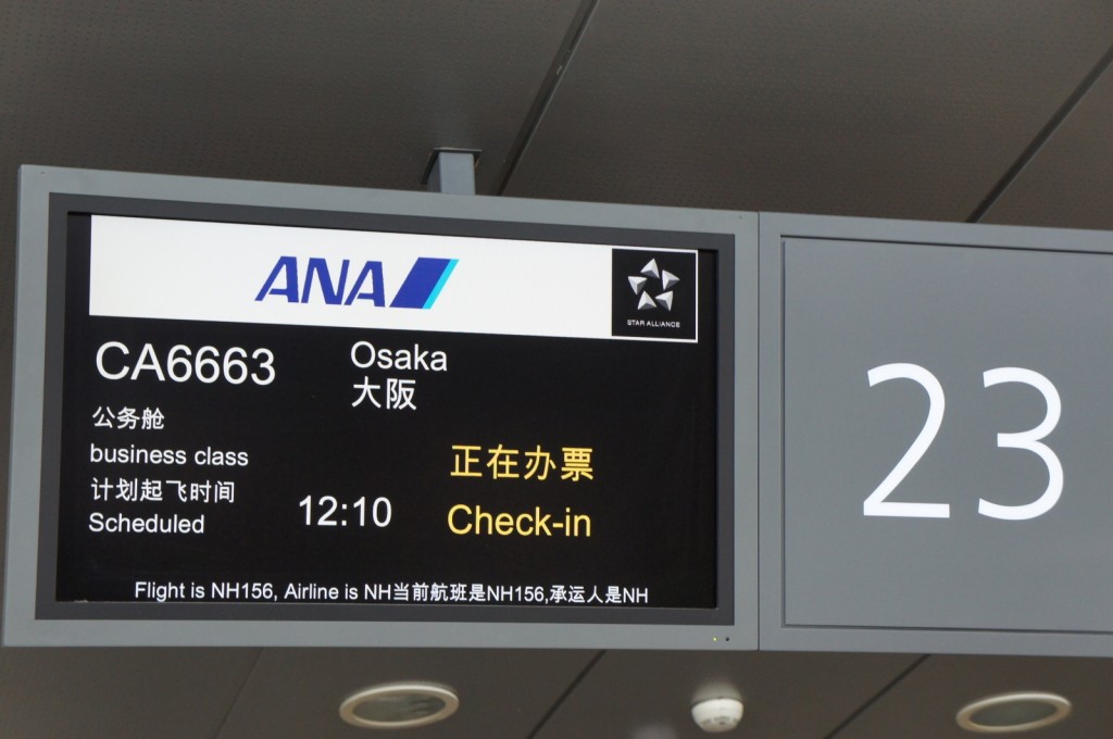 The image shows an airport departure information screen. The screen displays the following details:

- Airline: ANA (All Nippon Airways)
- Flight number: CA6663
- Destination: Osaka
- Class: Business class
- Scheduled departure time: 12:10
- Status: Check-in (indicated in both English and Chinese)
- Gate number: 23
- Additional information: "Flight is NH156, Airline is NH" (in both English and Chinese)
- The Star Alliance logo is also visible on the screen.
