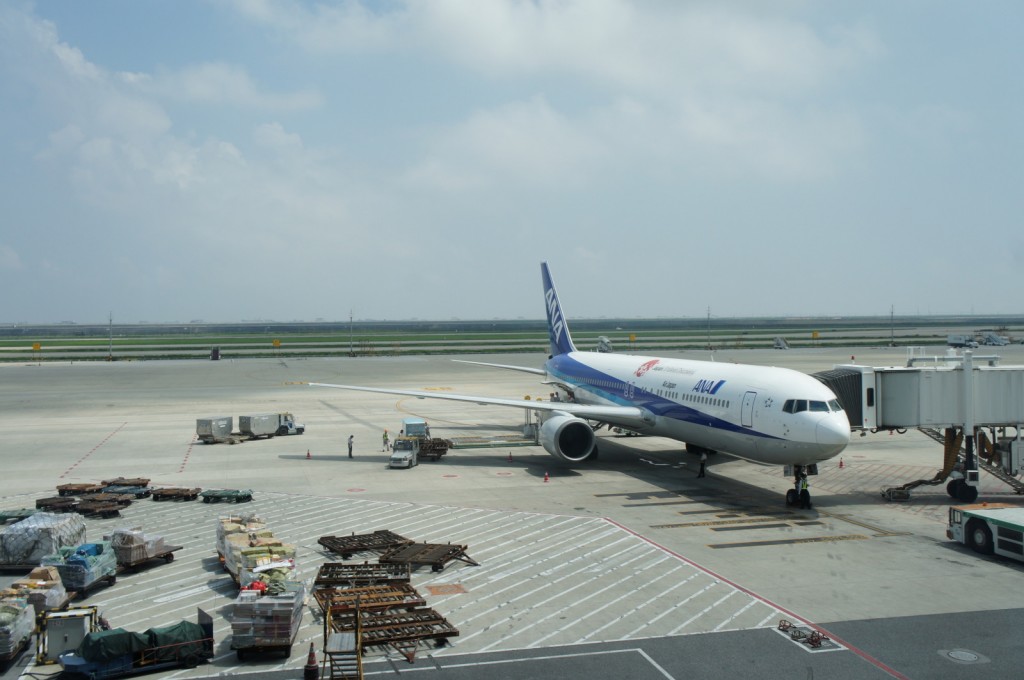 The image shows an ANA (All Nippon Airways) airplane parked at an airport gate. The aircraft is connected to a jet bridge, and there are various ground service vehicles and equipment around it. The sky is partly cloudy, and the runway and taxiway are visible in the background. There are also some cargo and luggage carts in the foreground.