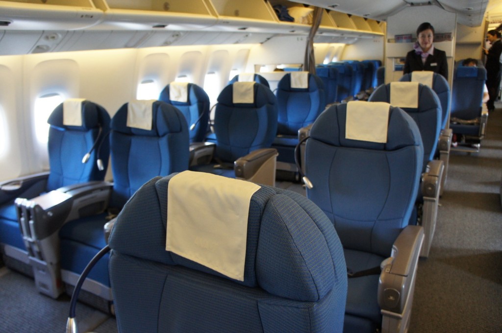 The image shows the interior of an airplane cabin, specifically the business or first-class section. The seats are large, blue, and well-cushioned, with white headrest covers. There are individual reading lights attached to the seats. Overhead compartments are visible above the seats. A flight attendant is standing in the aisle towards the back of the cabin, smiling. The cabin appears clean and well-lit.