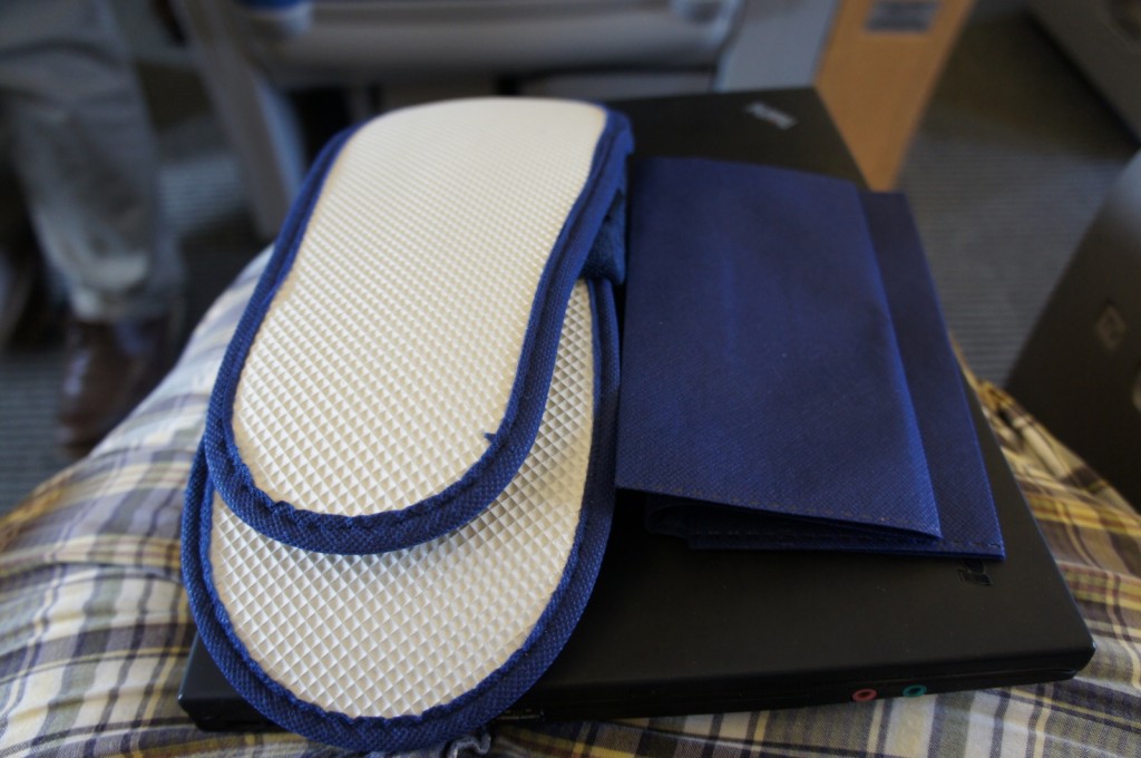The image shows a pair of white and blue slippers placed on top of a black laptop. Next to the slippers, there is a folded blue cloth or napkin. The items are resting on someone's lap, who is wearing plaid-patterned clothing. The background appears to be the interior of an airplane or a similar setting.