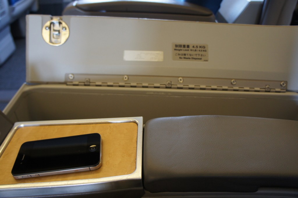 The image shows an open storage compartment on an airplane, with a weight limit of 4.5 kg indicated on a label inside the compartment. A smartphone is placed on a small tray next to the compartment. The interior of the airplane, including seats and other parts of the cabin, is visible in the background.