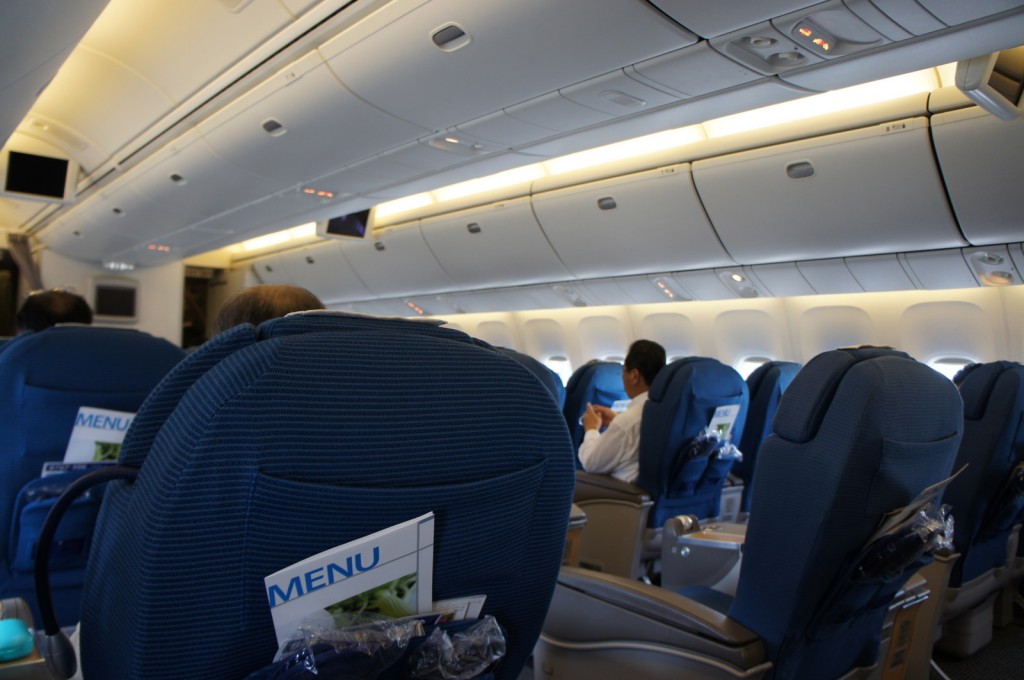 The image shows the interior of an airplane cabin. The seats are upholstered in blue fabric, and there are several passengers seated. Each seat has a pocket on the back containing a menu. The overhead compartments are closed, and the cabin lighting is on. Some small screens are visible on the ceiling, likely for in-flight entertainment. The overall atmosphere appears calm and orderly.