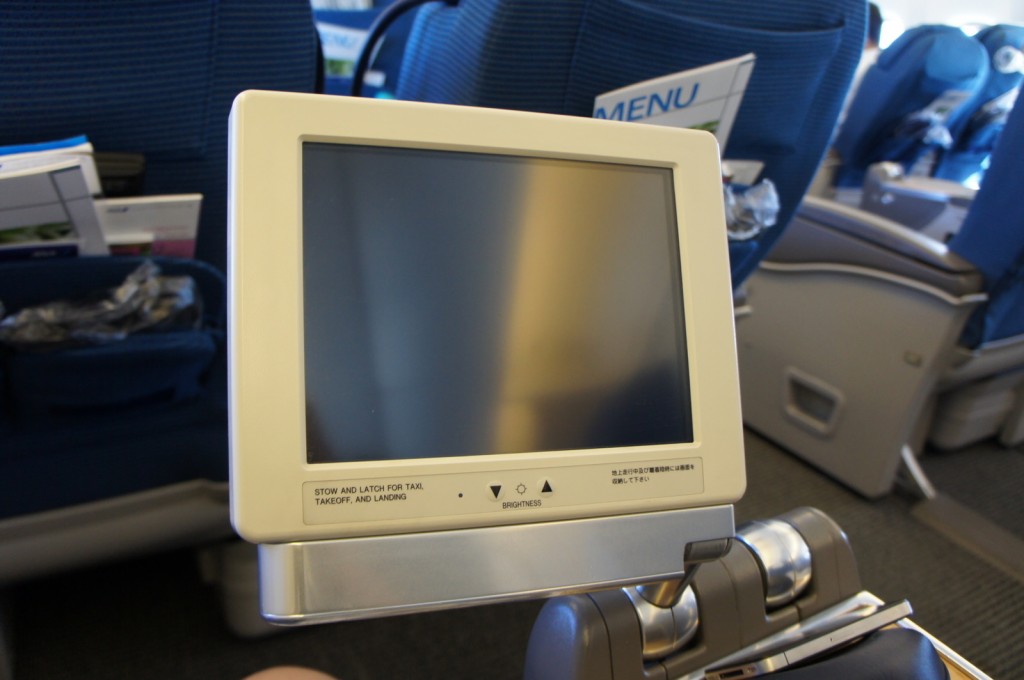 The image shows the interior of an airplane cabin, focusing on an in-flight entertainment screen attached to the back of a seat. The screen is turned off, and there is a label below it that reads "STOW AND LATCH FOR TAXI, TAKEOFF, AND LANDING." The background includes other airplane seats, some with seatback pockets containing magazines and other items. The overall setting suggests a commercial flight.
