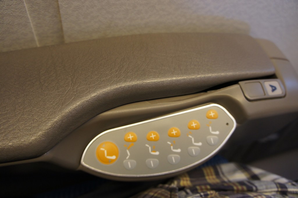 The image shows the armrest of an airplane seat with a control panel embedded in it. The control panel has several buttons, including ones with plus and minus signs, and icons indicating seat adjustments such as reclining and leg rest positions. The armrest is covered in brown leather or leather-like material.