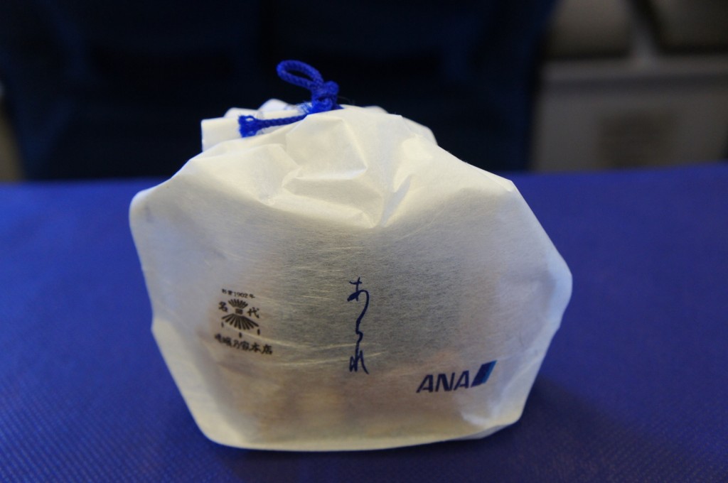 A small package wrapped in white paper with blue string on top, placed on a blue surface. The package has Japanese text and logos printed on it, including the ANA logo. The background is slightly blurred, suggesting the image might have been taken in an airplane or similar setting.
